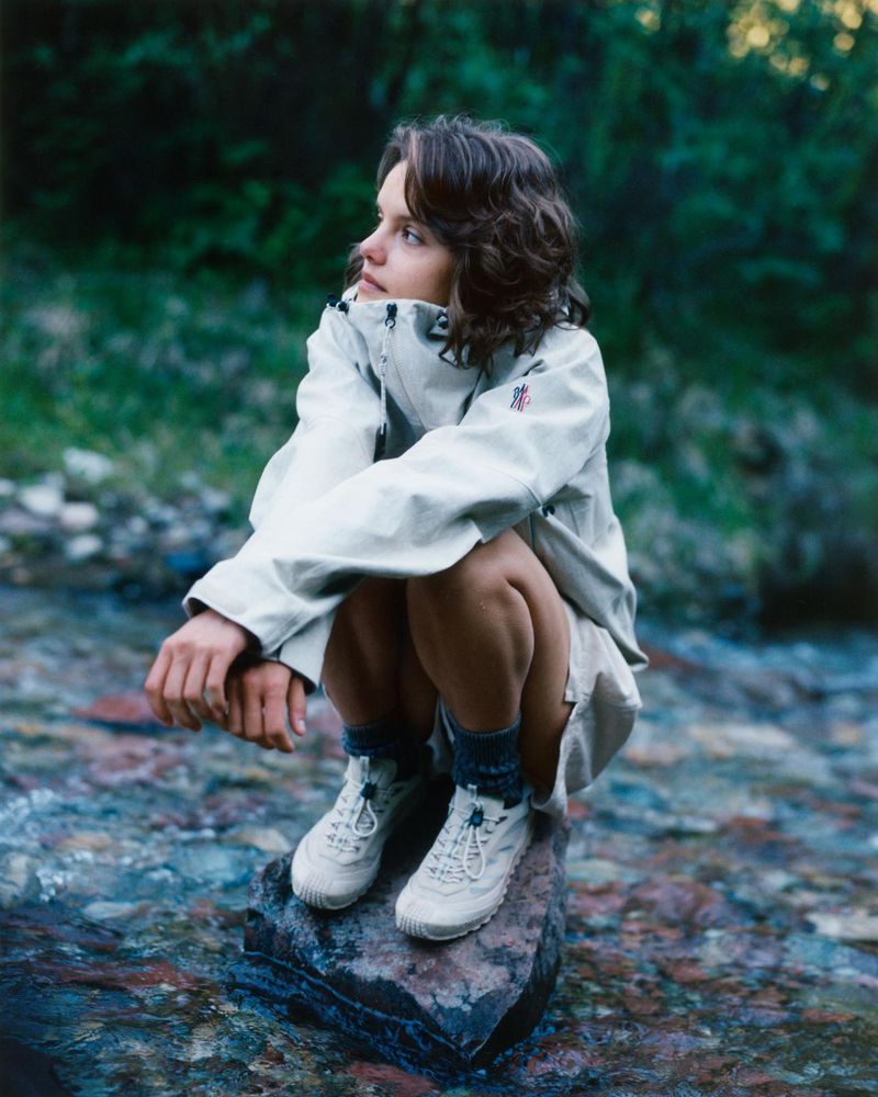 Woman crouching on a rock in a shallow stream wearing an off-white technical jacket and trail sneakers.