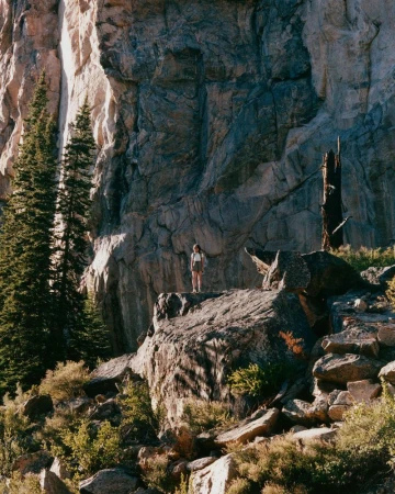 A lone model stands atop a rugged rock formation framed by towering granite cliff faces and evergreen pine trees.