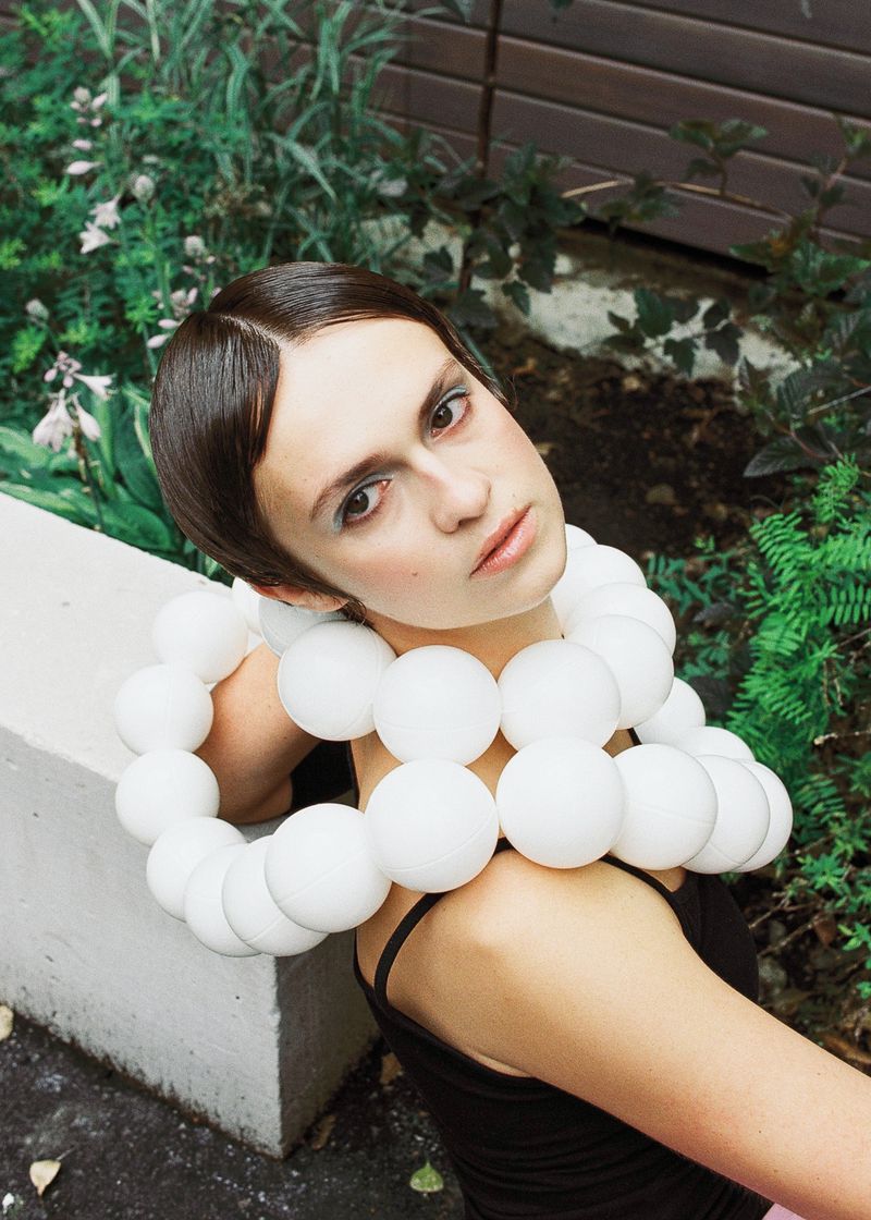 Woman with short dark hair wearing a large sculptural white spherical beaded necklace and black top outdoors next to garden foliage.