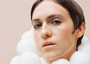Close-up portrait of a person with light blue eyeshadow wearing a sculptural collar of white plastic spheres against a pale backdrop.