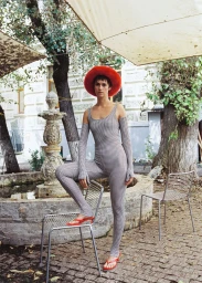 Model wearing a checkered catsuit, wide-brim orange-red hat, and red heels resting one foot on a metal mesh chair outdoors by a fountain.
