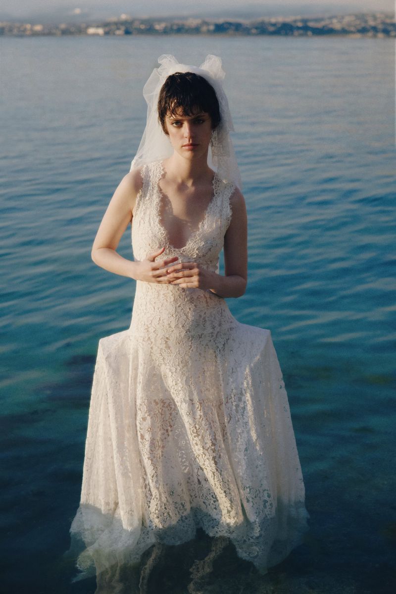 A woman with short dark hair wears a white lace wedding dress and veil while standing in the shallow blue water of a lake or sea.