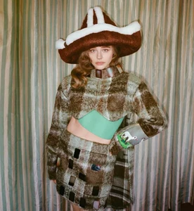 Chloe Grace Moretz wearing an oversized brown and white plush hat and a fuzzy plaid two-piece outfit against striped curtains.