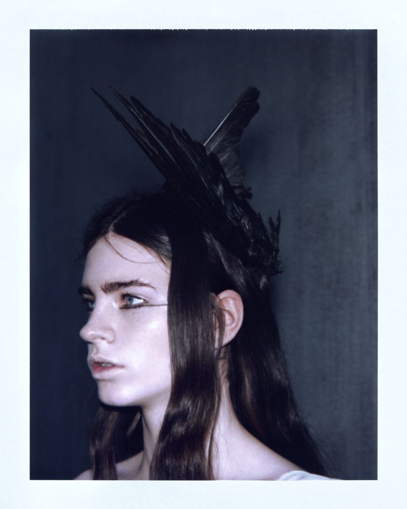 Profile portrait of a woman wearing an avant-garde black feather headpiece and graphic eyeliner against a dark backdrop.
