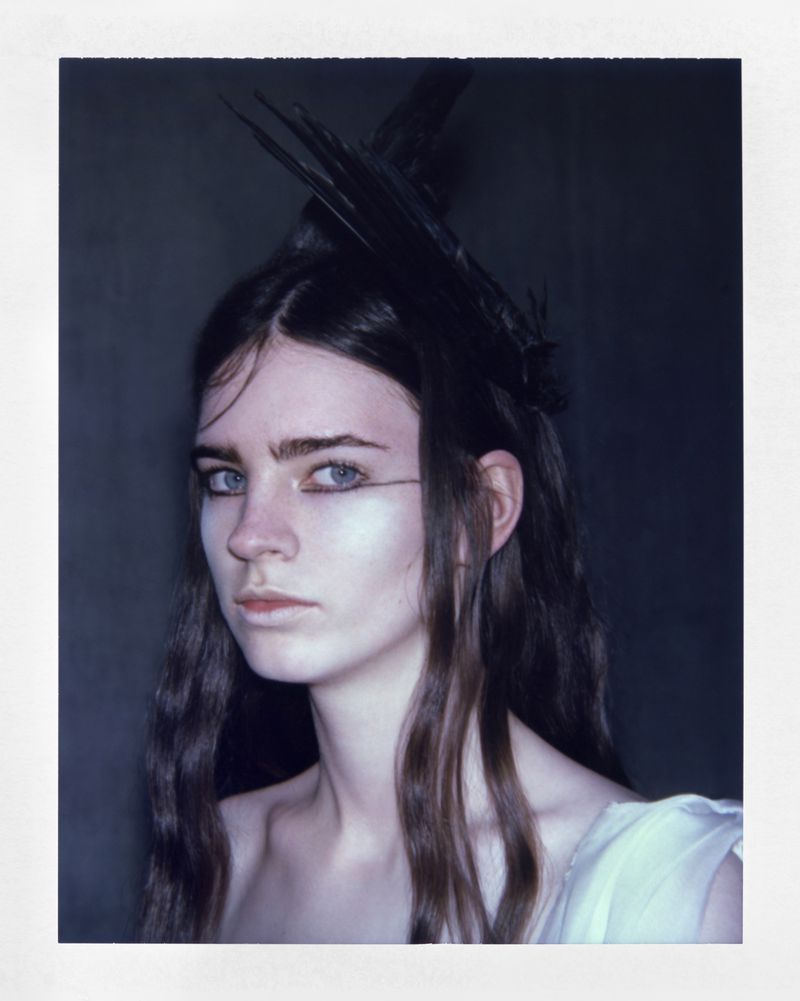 Close-up portrait of a woman with long dark hair wearing a spiky black feather headpiece and white garment against a dark backdrop.