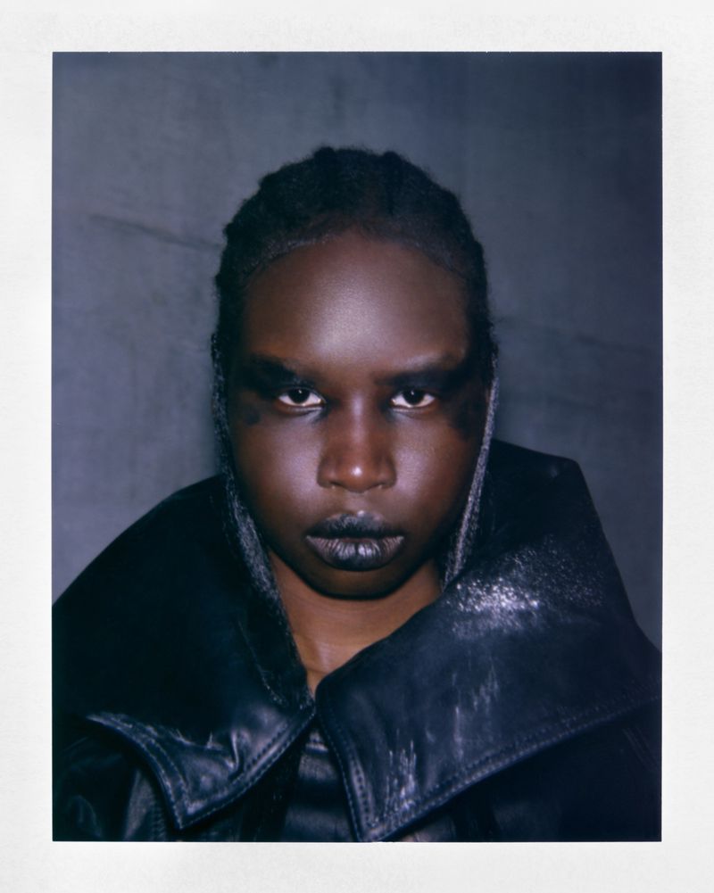 A close-up portrait of a dark-skinned model with bold black eye makeup and dark lips, wearing a structured black leather-texture jacket.