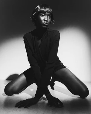 Black and white portrait of a black woman crouching on the floor wearing a dark top, with dramatic lighting creating deep shadows.