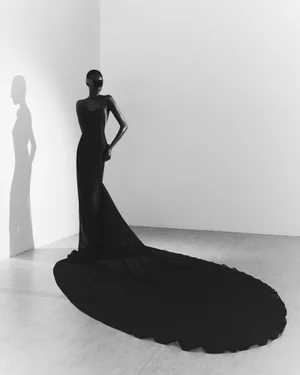 Model Aluk Malual wearing a long black gown with a train, casting a sharp shadow against a plain white studio wall.