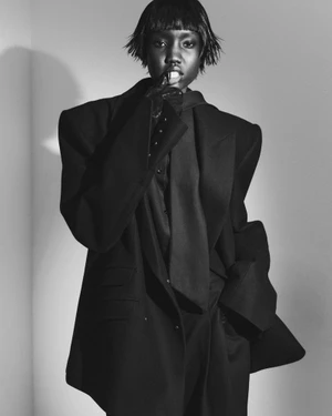 Monochrome fashion editorial portrait of a model in a structured black blazer with sharp padded shoulders against a plain background.