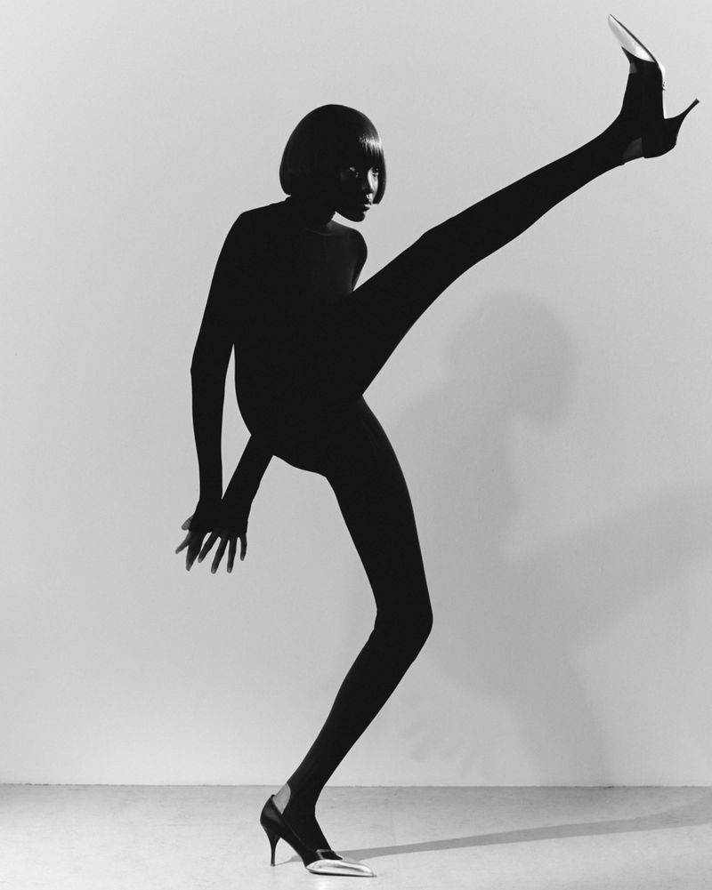 Black and white studio shot of a slender adult woman with a dark bob haircut in a catsuit kicking her leg up wearing pointed-toe heels.