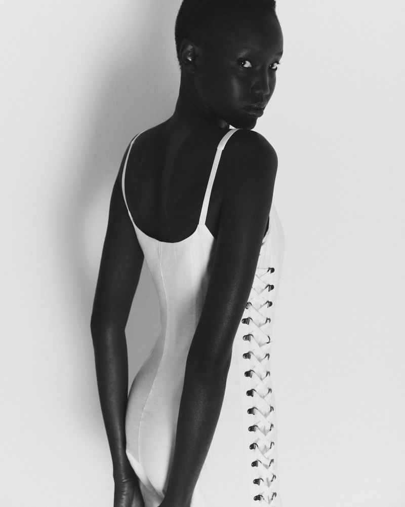 Woman in white dress poses against a white background. Portrait shot shows bare arms and back with lace-up dress detail.