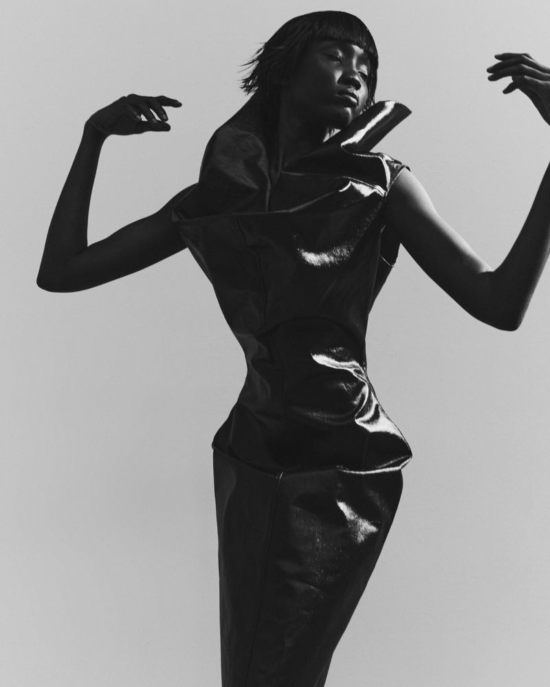 Monochrome fashion shot of an African American woman in a sculpted shiny dress with raised arms against a plain background.