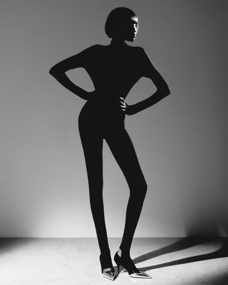 Full-body silhouette of an adult female fashion model in a catsuit, wearing high heels, posing in a photo studio against a grey background.