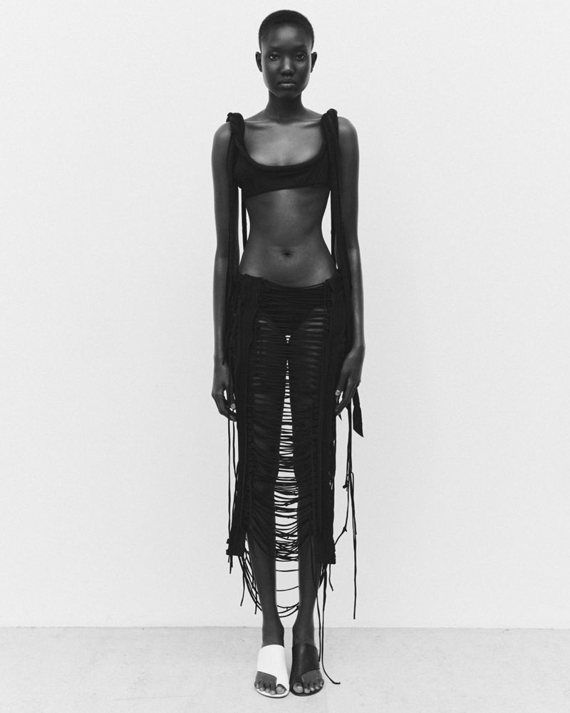 Full-length shot of an African female fashion model posing in a black bra and skirt with fringe details against a white background.