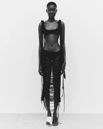 Full-length shot of an African female fashion model posing in a black bra and skirt with fringe details against a white background.