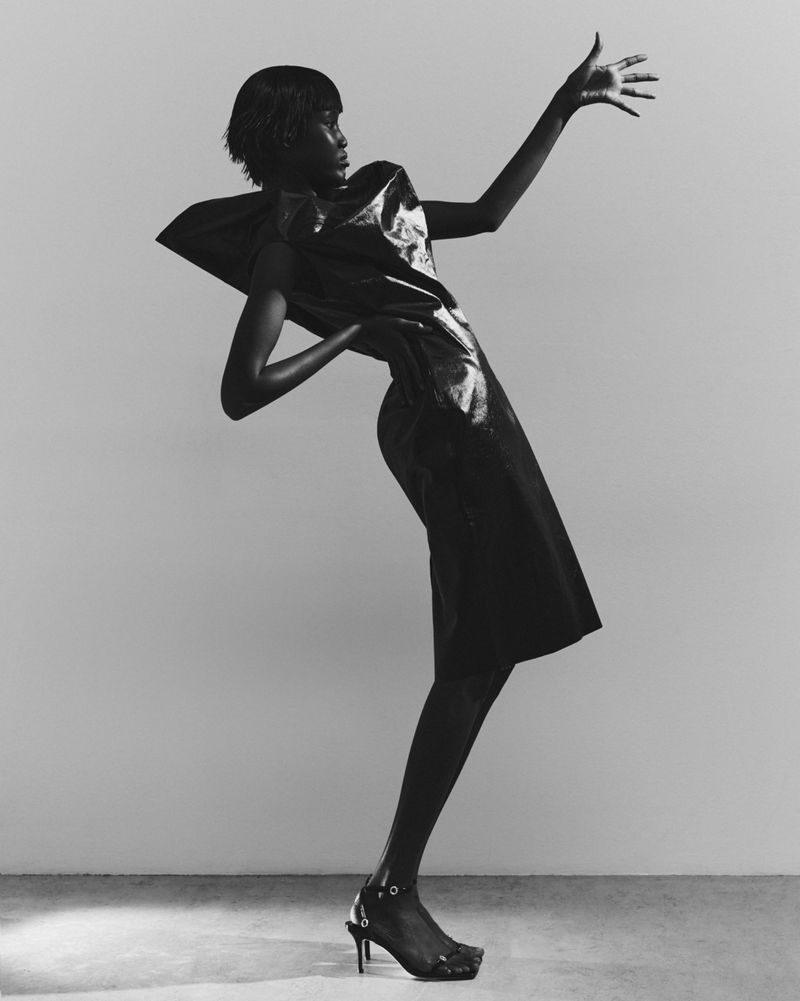 Monochrome fashion shot of a Black woman in a structured dress and heels, posing with hand raised against a neutral backdrop in a photo studio.