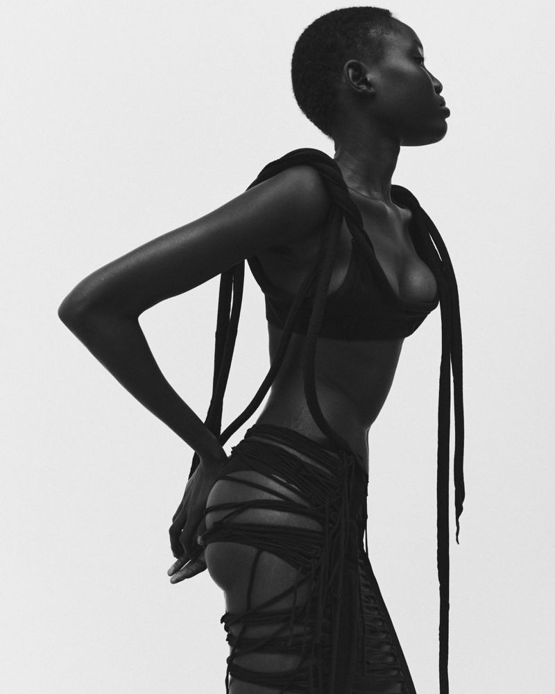 Striking black and white portrait of an African woman in distinctive dark, stringy fashion against a clean, light background.
