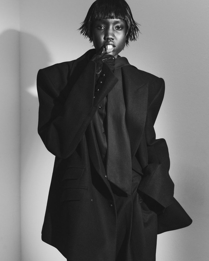 Monochrome fashion portrait of a Black woman with short hair wearing an oversized dark blazer, posing in a corner studio setting.