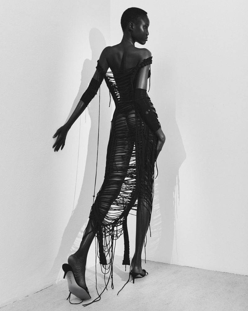 Black and white fashion shot of a slender adult female model in an open-backed dress and high heels, posing in a minimalist corner.