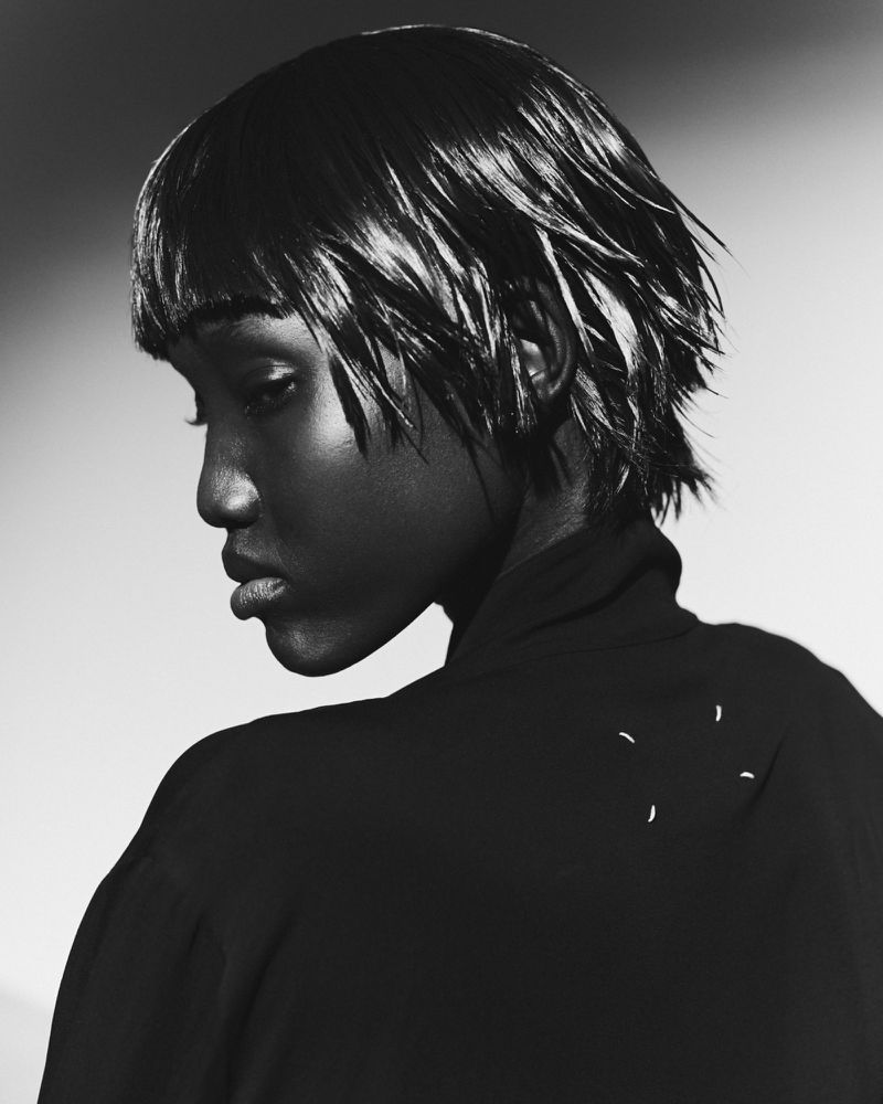 Black and white portrait of a Black woman with short, wet hair, wearing a dark top, posing in a photo studio with a light background.