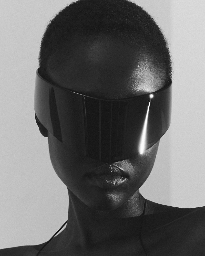 Close-up studio shot of a Black woman wearing a futuristic visor, showcasing glossy black material and neutral skin tone against a white backdrop.