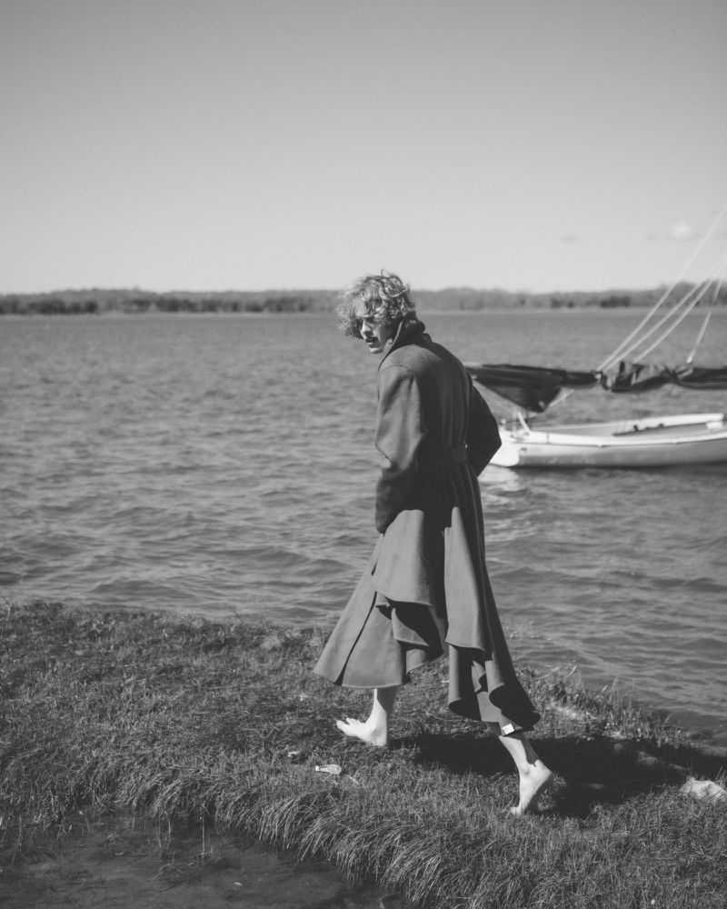 Barefoot person with curly hair walking along a grassy shoreline wearing a long draped coat in black and white.