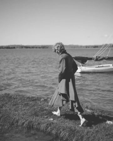 Barefoot person with curly hair walking along a grassy shoreline wearing a long draped coat in black and white.