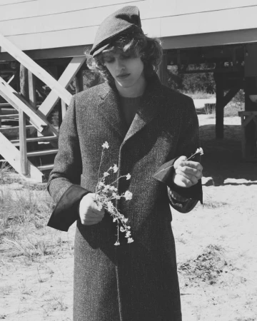 A male model wearing a dark textured overcoat and beret holds small wildflowers outdoors in a black and white fashion editorial.