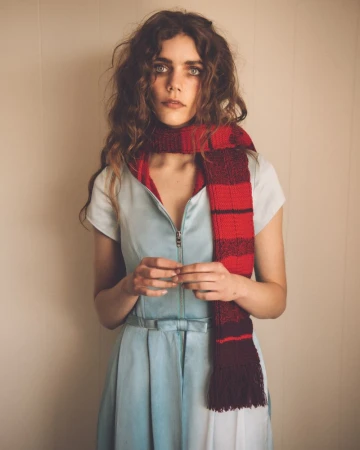 Woman with long wavy hair wearing a light blue zip-front dress and a red striped knitted scarf against a neutral wall.