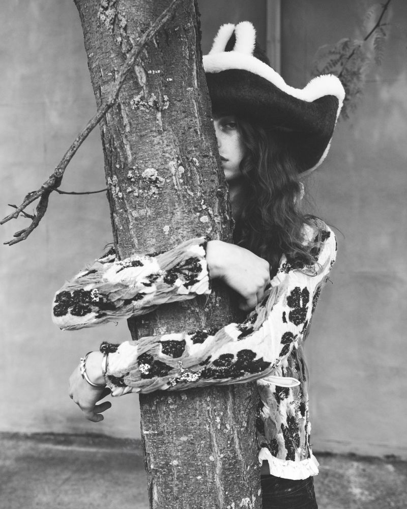 A model wearing a plush-trimmed tricorn hat and patterned jacket hugs a tree trunk in a black and white fashion portrait.