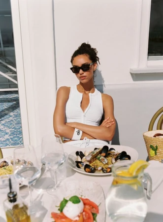 Woman in dark sunglasses and white Alo activewear seated with arms crossed at a sunlit outdoor dining table.