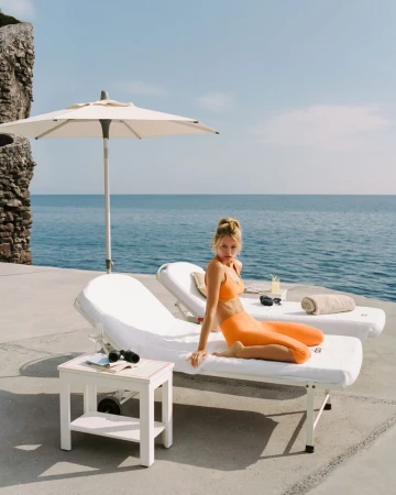 Model Frida Aasen kneeling on a white sun lounger overlooking the ocean, wearing an orange activewear set for Alo.