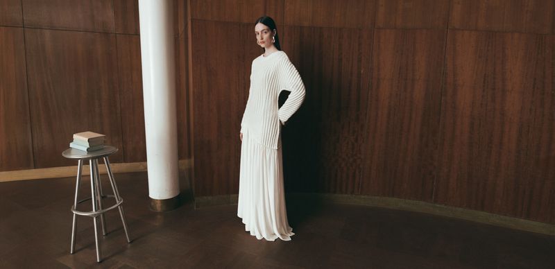 Model Lila Braghero in a long ivory pleated knit dress standing beside a wooden wall and pillar next to a silver stool.