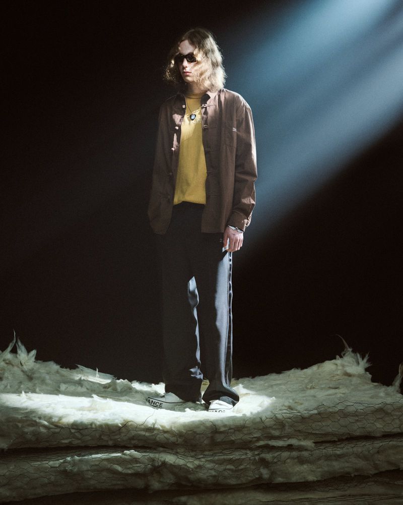 Model in dark sunglasses, a brown overshirt, yellow t-shirt, and black wide-leg trousers standing in a spotlight on a textured studio set.