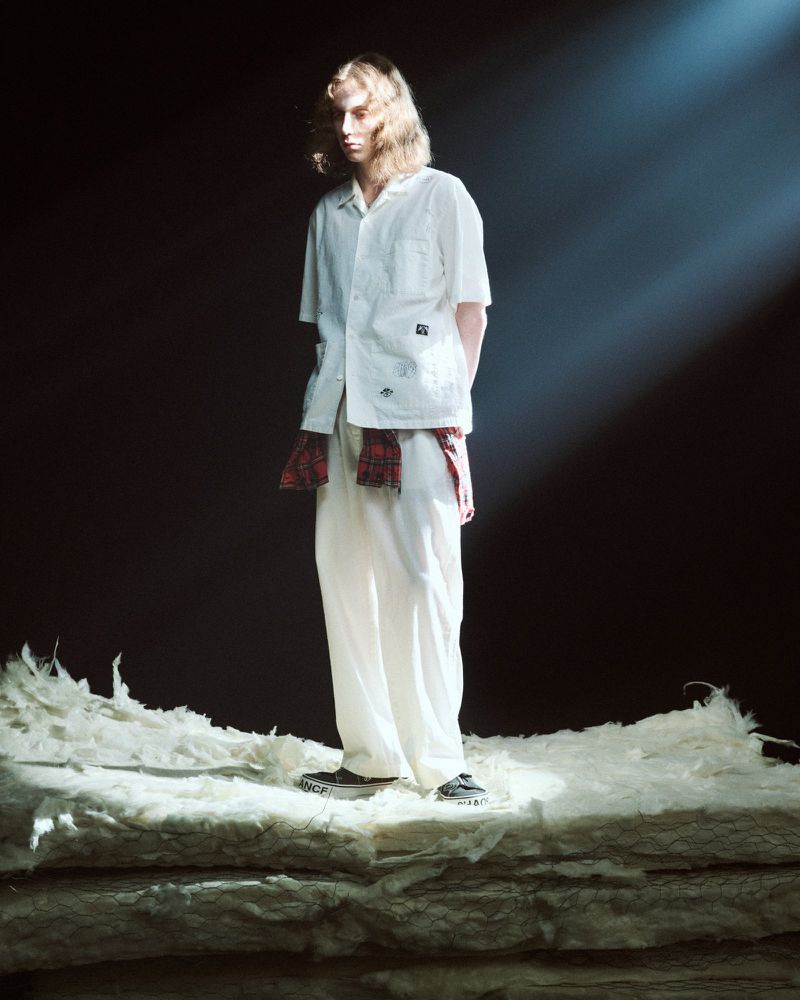 Model standing under a diagonal spotlight wearing an off-white short-sleeve shirt, matching trousers, and a plaid waist tie.