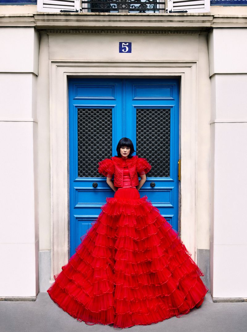 Woman in a voluminous red ruffled gown stands in front of a blue door with a number 5 sign, creating a striking contrast.