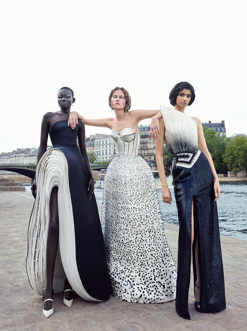 Three female models pose in elegant couture gowns along a stone walkway with a city and river in the background on an overcast day.