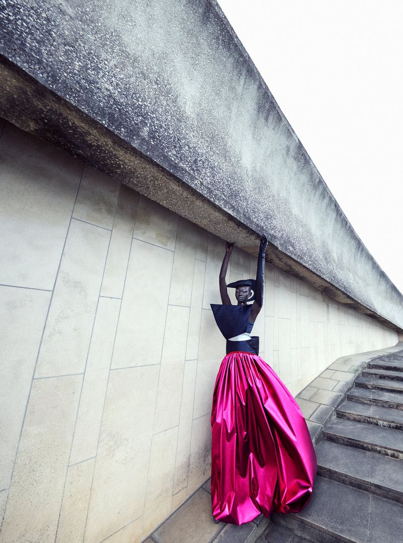 Fashionable woman in artistic outdoor scene. She wears a black top and shiny pink skirt, posing under concrete architectural element.