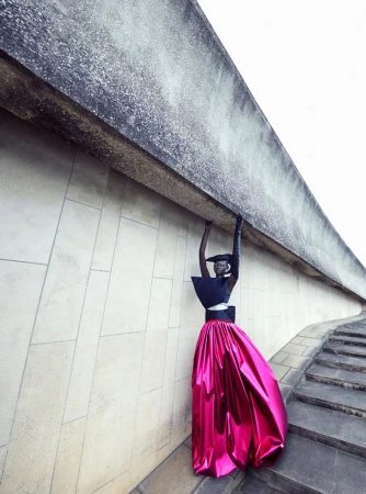 Fashionable woman in artistic outdoor scene. She wears a black top and shiny pink skirt, posing under concrete architectural element.