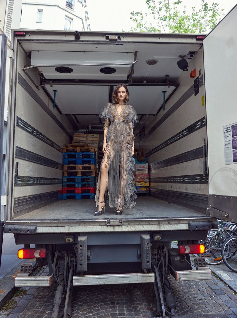 A young woman in an elegant sheer gown stands in the open back of a box truck, her high heels planted on the truck's loading platform.