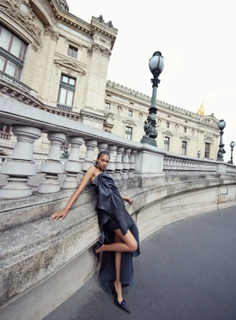 Fashion editorial image of an adult female model in a black strapless dress posing on a bridge with ornate architecture in the background.