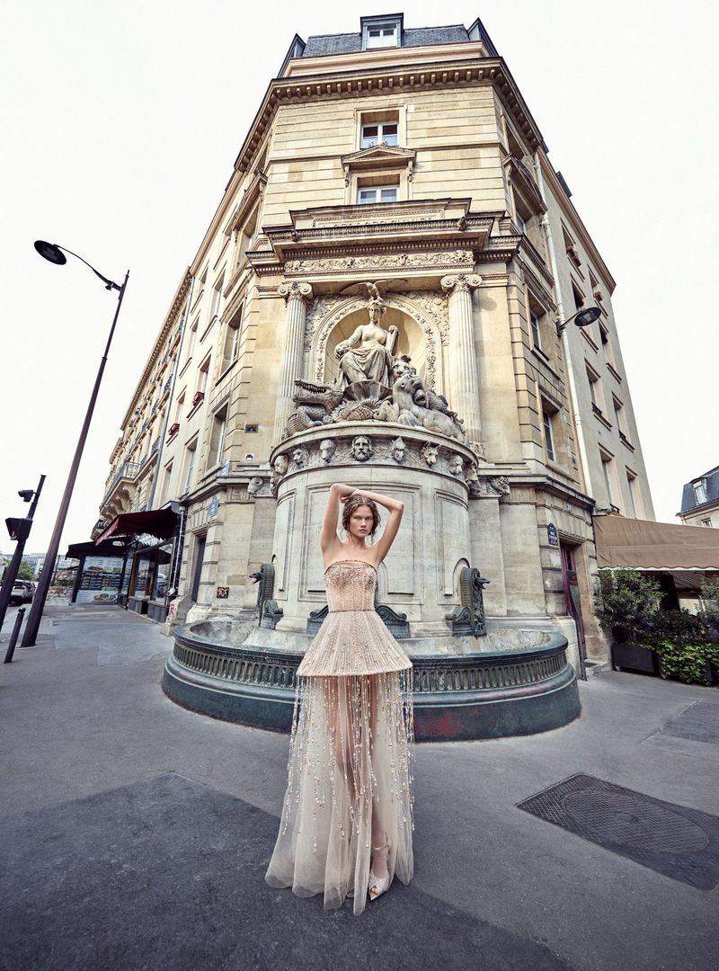 Fashion shot of a model in an ornate gown posing in front of a historic building with a fountain in an urban setting with neutral tones.
