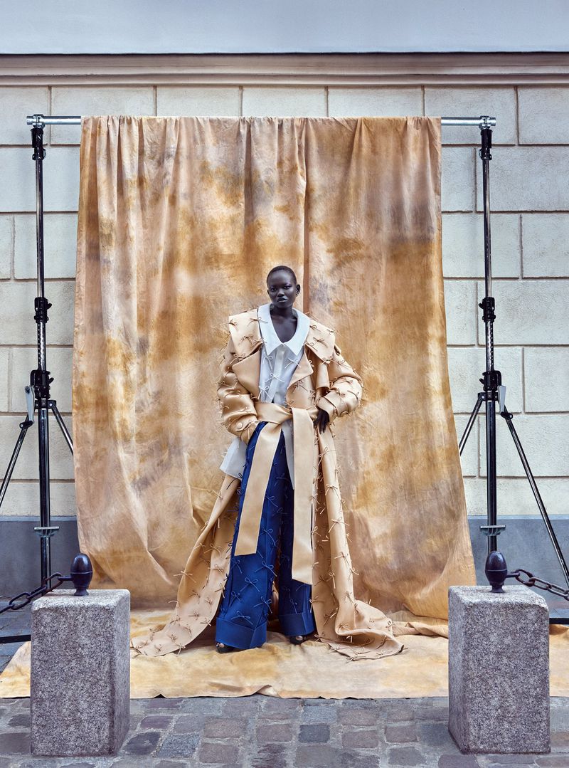 Fashion portrait of a woman in a trench coat and wide-leg pants standing in front of a beige backdrop and metal light stands.