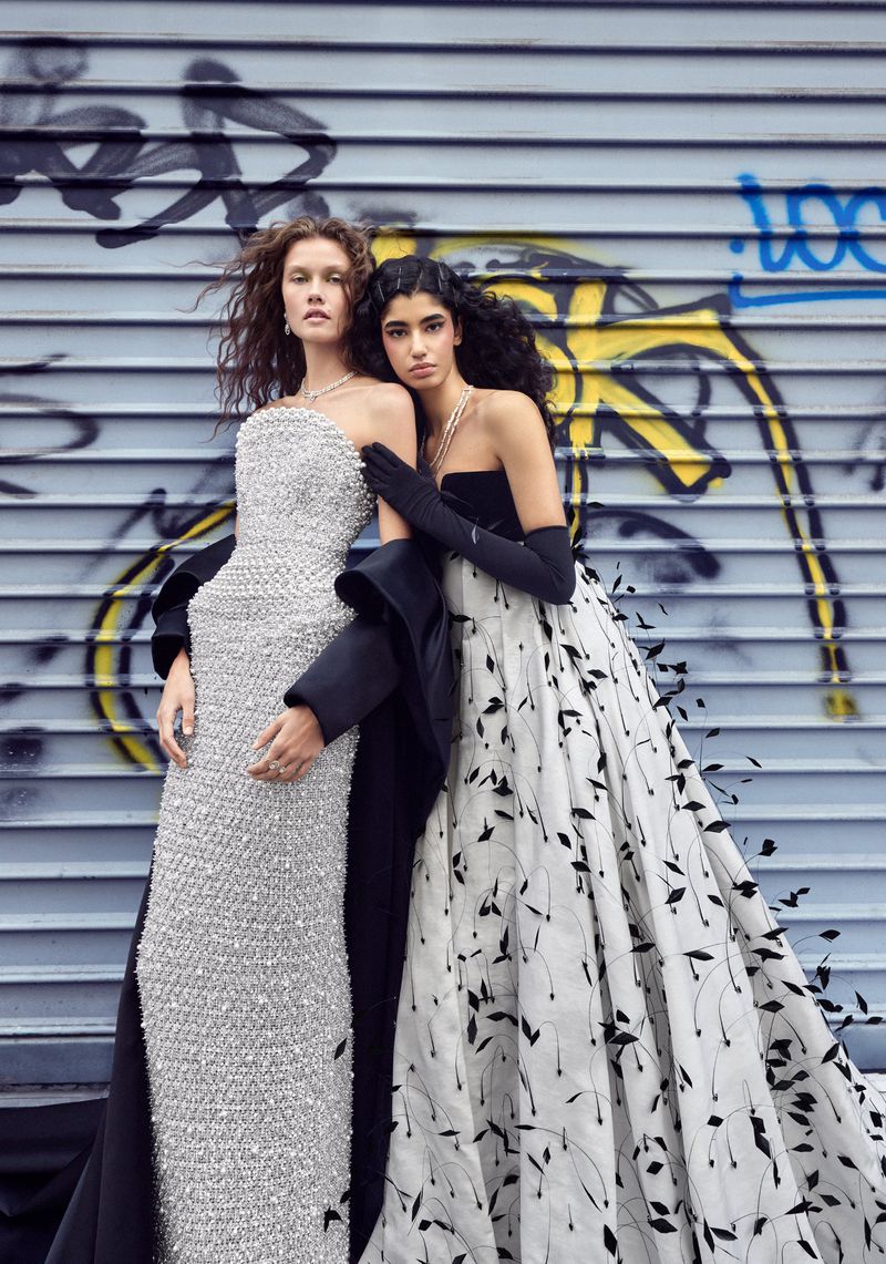 Two women pose in elegant evening gowns against a graffiti-covered wall, one in silver sequins, the other in black and white. Fashion editorial.