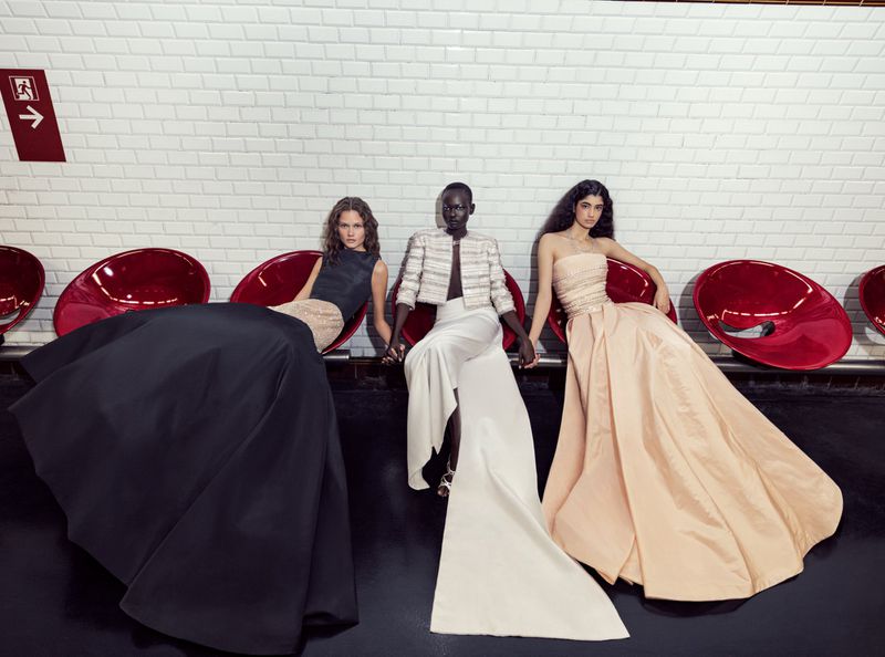 Three women in elegant evening gowns sit in a subway station, showcasing high fashion with a modern and sophisticated aesthetic.