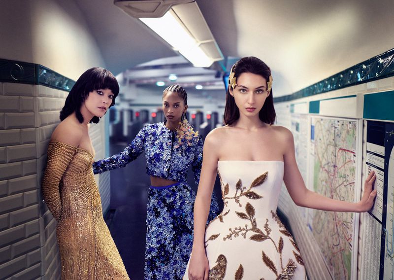 Three female fashion models pose in a subway, wearing designer dresses in white, gold, and blue, showcasing elegant style and beauty.