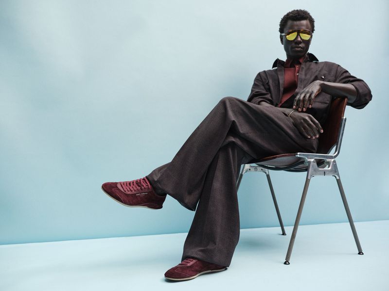 Black man seated on a chair wearing a dark tailored suit, yellow sunglasses, and burgundy sneakers against a blue background.