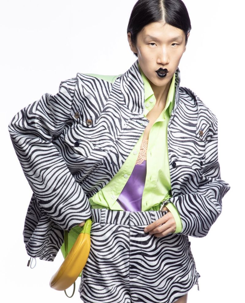 Model wearing a zebra print jacket and shorts set with a lime green shirt and holding a banana bag against a white studio backdrop.