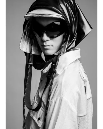 Black and white portrait of an Asian male model wearing dark visor sunglasses and an avant-garde technical hooded cap with hanging straps.