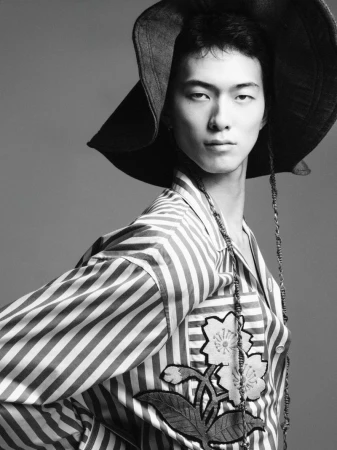 East Asian male model wearing a wide-brim hat and striped embroidered shirt in a black and white studio portrait.